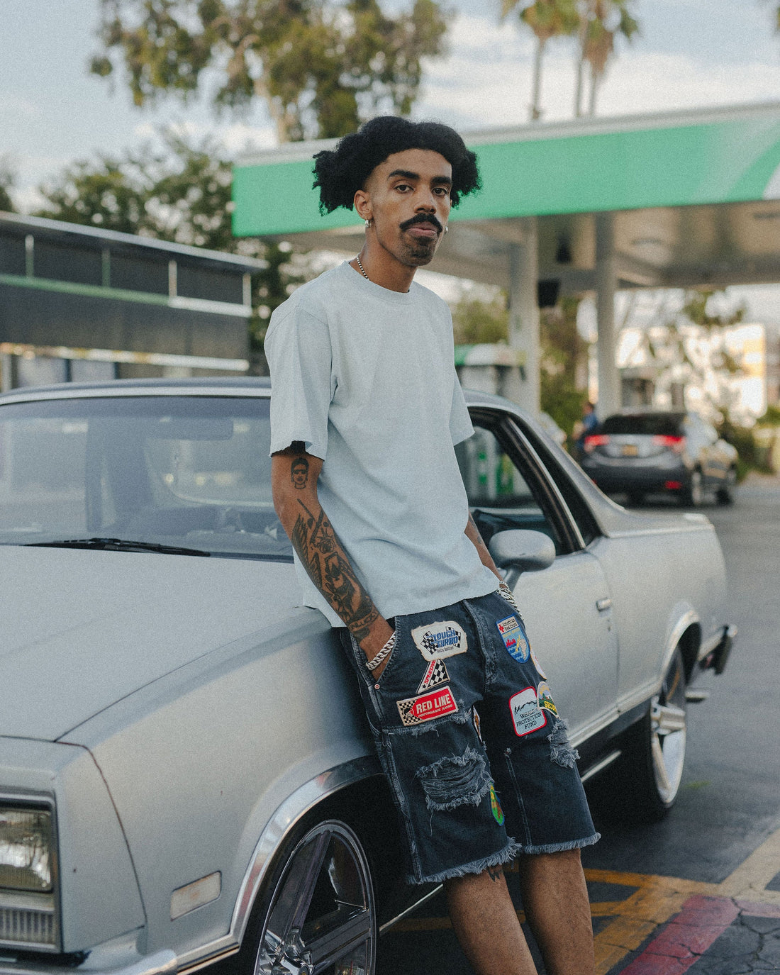  Model in light blue logo tshirt and patched denim shorts leaning against vintage car